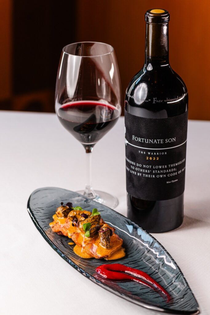 Hundred Acre Fortunate Son wine bottle beside a glass of red wine and a plated dish.