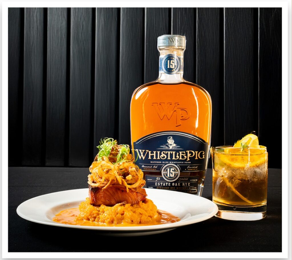 WhistlePig whiskey bottle displayed with a plated dish and whiskey cocktail